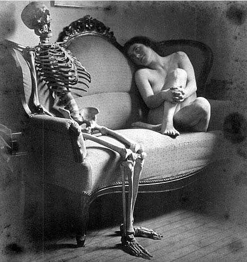 A nude woman sits on a couch looking at a skeleton