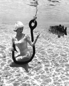 1950s photo of a woman in a bathing suit underwater sitting on a hook beckoning to a fish