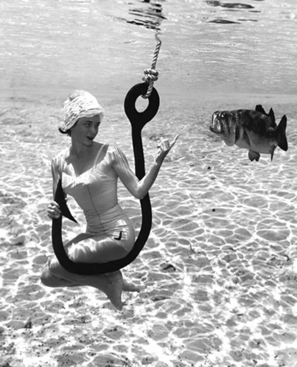 1950s photo of a woman in a bathing suit underwater sitting on a hook beckoning to a fish