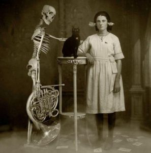 Vintage photo of a woman leaning on table with a black cat standing next to a skeleton holding a trombone.