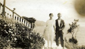 Image shows a bride and groom standing outside. A child can be seen watching the photograph be taken from steps on the left hand side of the image.