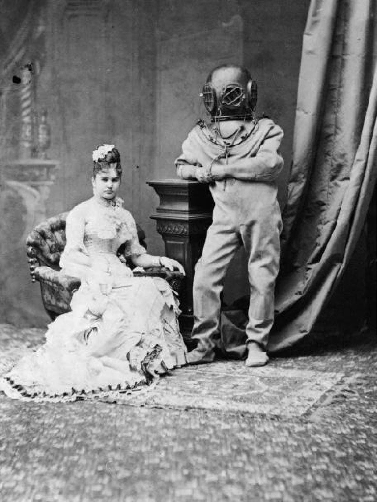 Vintage photo of people posing in a parlor. A woman seated in a white dress and a figure in an old fashioned scuba suit. 