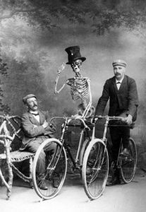 Vintage photo of a man on a bench looking at another man and a skeleton riding bikes