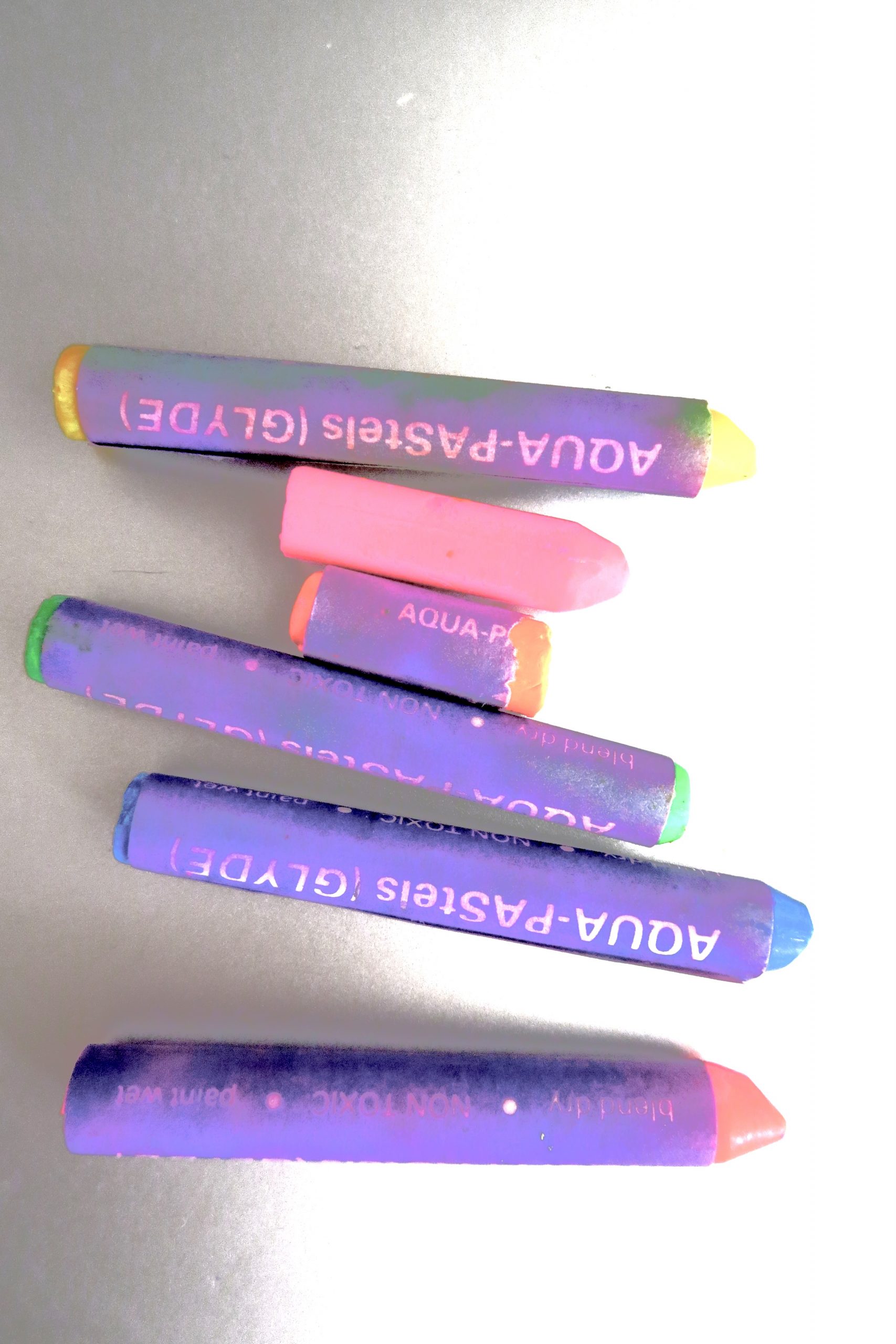 Five AQUA-PAStels (Glyde) water-soluble pastel crayons in purple, pink, green, and blue, arranged diagonally on a gray surface, with their labels showing in reverse orientation.