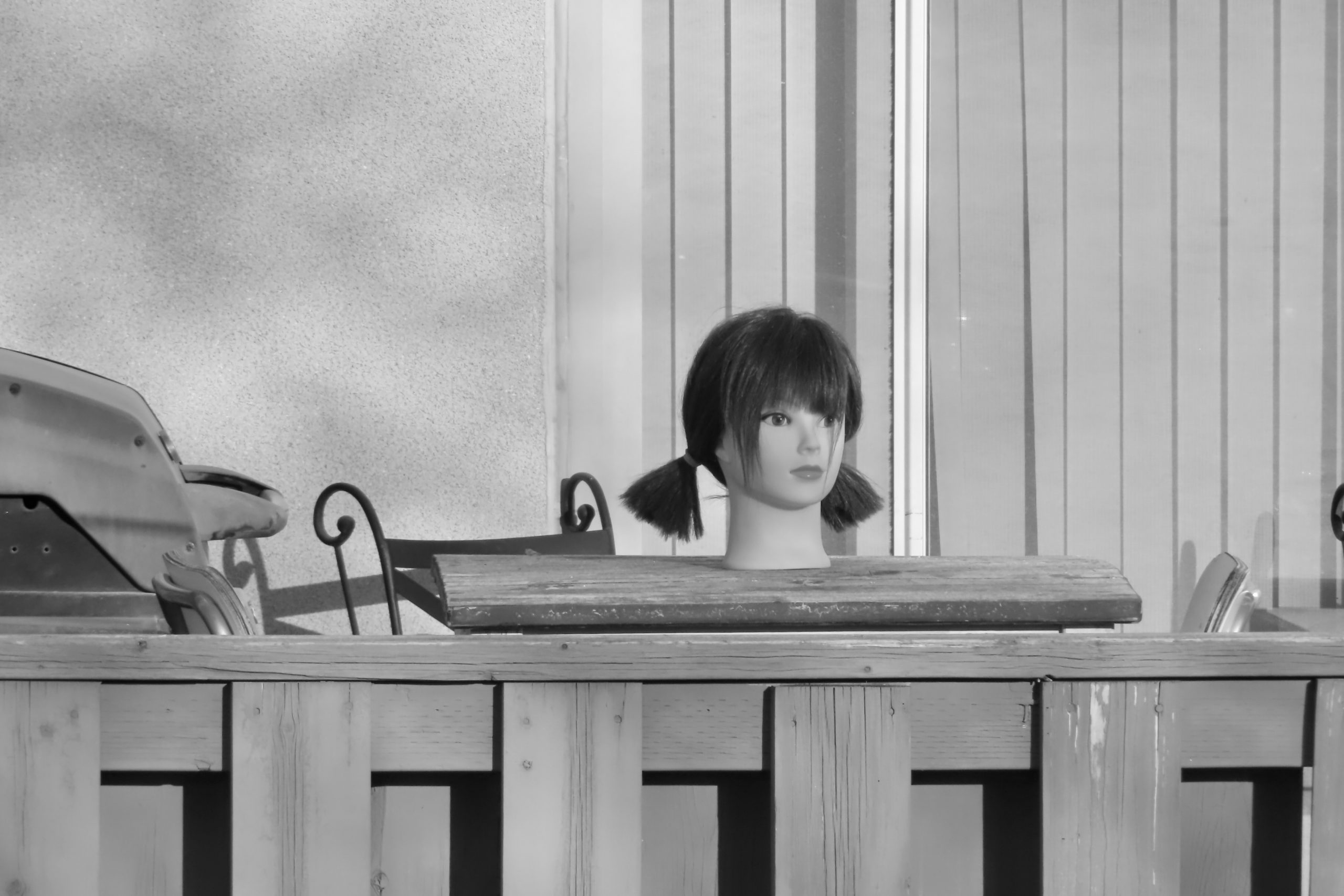 Black-and-white photograph of a mannequin head with short dark hair styled in two small pigtails, resting on a wooden surface. A corrugated metal wall fills the background, with metal chairs visible on either side. The scene has an industrial, workshop-like feel.