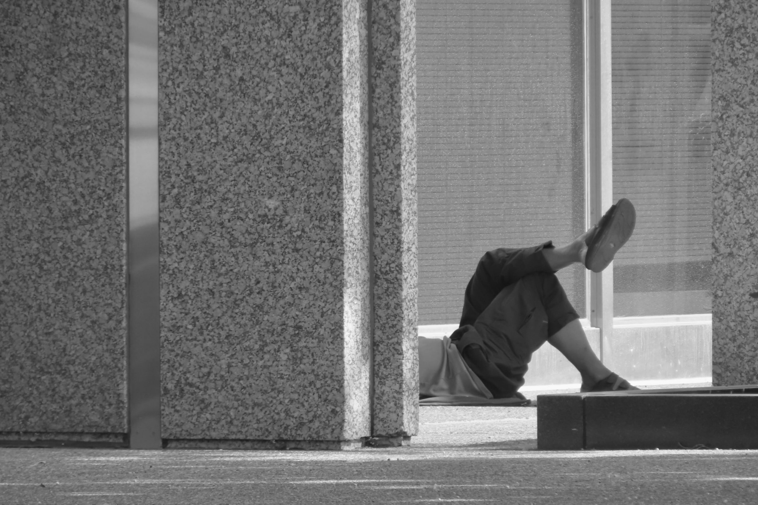 Black and white photo of a person in black shorts and sandles laying on the ground partially obscured by a building.