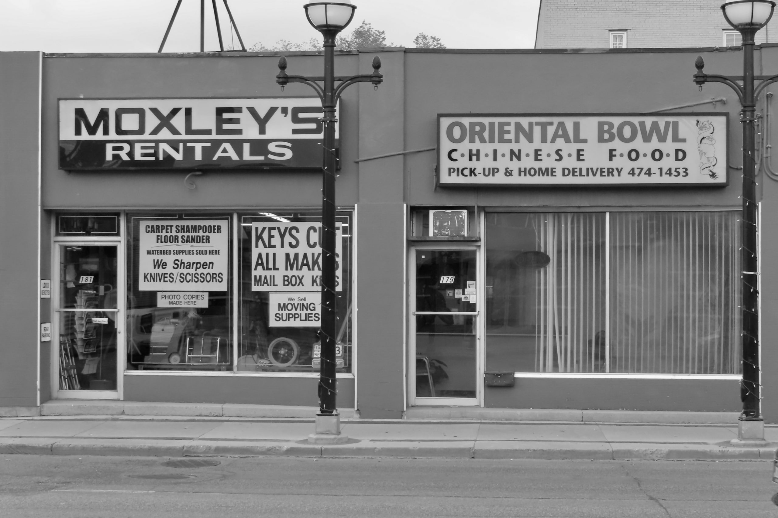 Black-and-white street-level photograph of a divided storefront. On the left, “Moxley’s Rentals” advertises carpet shampooers, floor sanders, knife sharpening, and photo copies. On the right, “Oriental Bowl Chinese Food” offers pickup and delivery. Signs for key cutting and moving supplies fill the windows. A street lamp and sidewalk anchor the foreground.