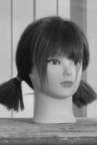 Black-and-white close-up photo of a mannequin head in profile with dark hair in pigtails and bangs, resting on a weathered wooden surface.