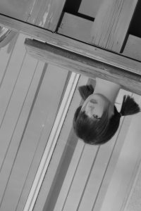 Black-and-white photo of a person’s upside-down reflection in glass, showing dark hair in pigtails, framed by a wooden border against a background of vertical slats.