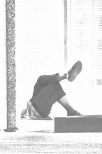Black-and-white photo of a person lying on their back with legs raised in the air, silhouetted against a bright, sunlit window with blinds.