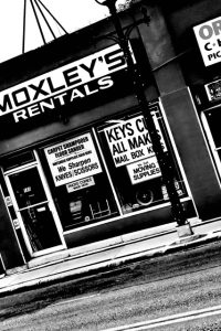 Dutch angle Black-and-white photo of the “MOXLEY’S RENTALS” storefront with large display windows advertising rentals, key cutting, and moving supplies, with a classic lamppost on the sidewalk.
