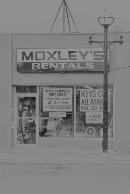 IMG_9723 Black-and-white photo of the “MOXLEY’S RENTALS” storefront with large display windows advertising rentals, key cutting, and moving supplies, with a classic lamppost on the sidewalk.