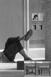 Black-and-white photo of a person sitting on a low wall outside a storefront, with a reflection of a woman visible in the glass window above and a small inset photo of two street shops in the corner.